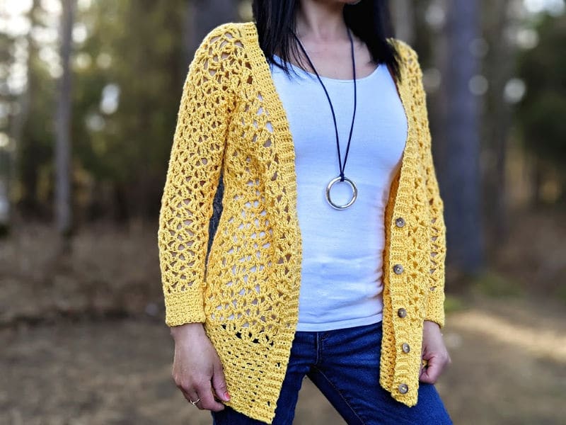 The Addy Lace Cardigan in a bright yellow is worn over a white tank top and jeans. The model is standing in front of a wooded area.