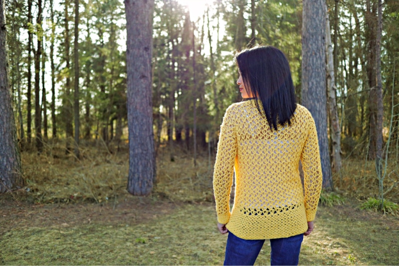 The Addy Lace Cardigan in a bright yellow is worn over a white tank top and jeans. The model is standing in front of a wooded area with her back facing the camera.