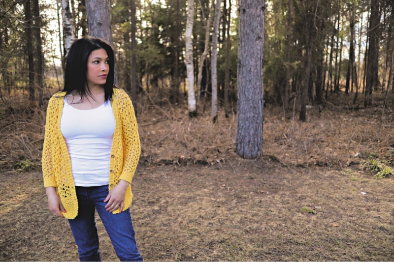 The Addy Lace Cardigan in a bright yellow is worn with the buttons undone, over a white tank top and jeans. The model is standing in front of a wooded area looking off to her left.