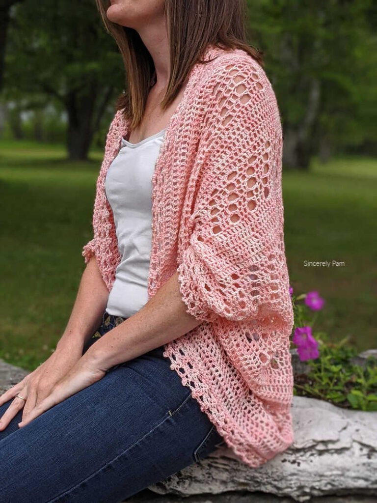 Model is sitting on a rock in a garden while wearing the Daphne cocoon cardigan crochet pattern by Sincerely Pam