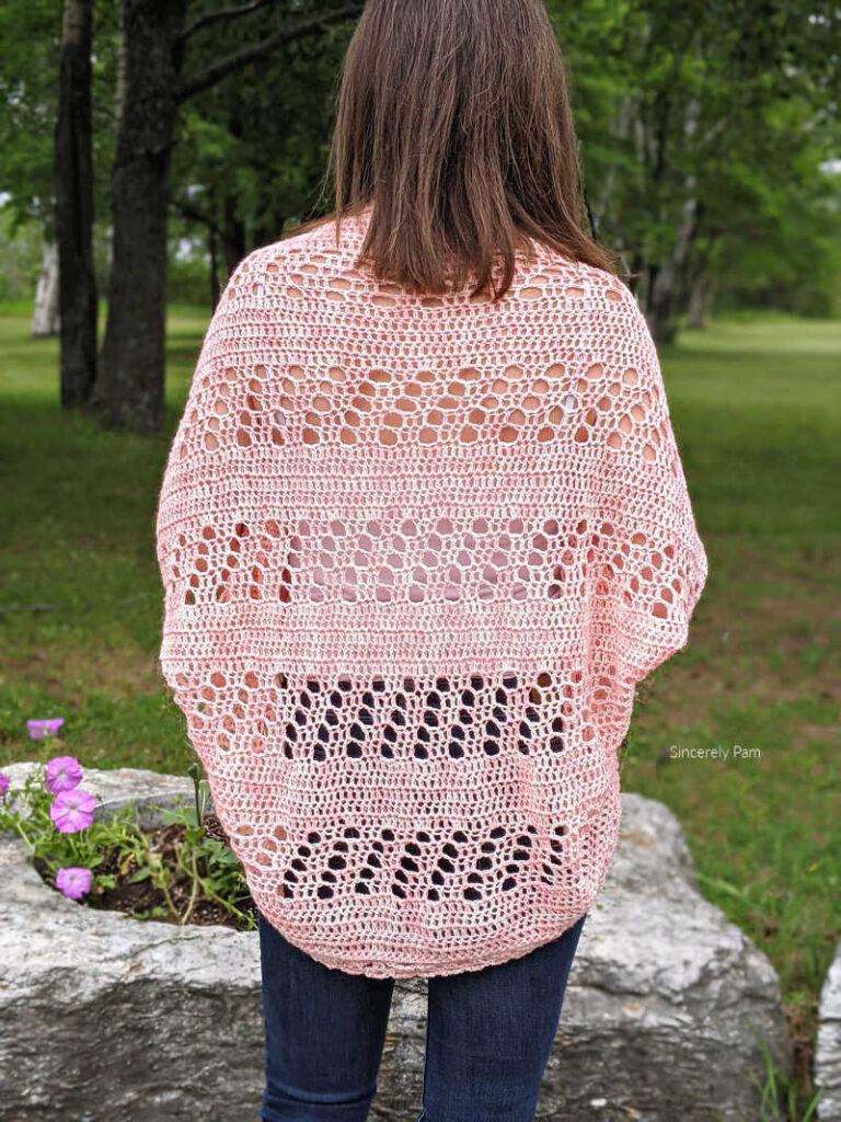 model is standing in a garden with her back to the camera. The daphne cocoon cardigan shows a filet crochet back detail.