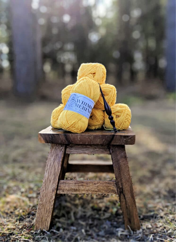 A stack of Whims Merino DK yarn in mustard and an Alpha Hook sit on top of a wooden stool on the grass. 