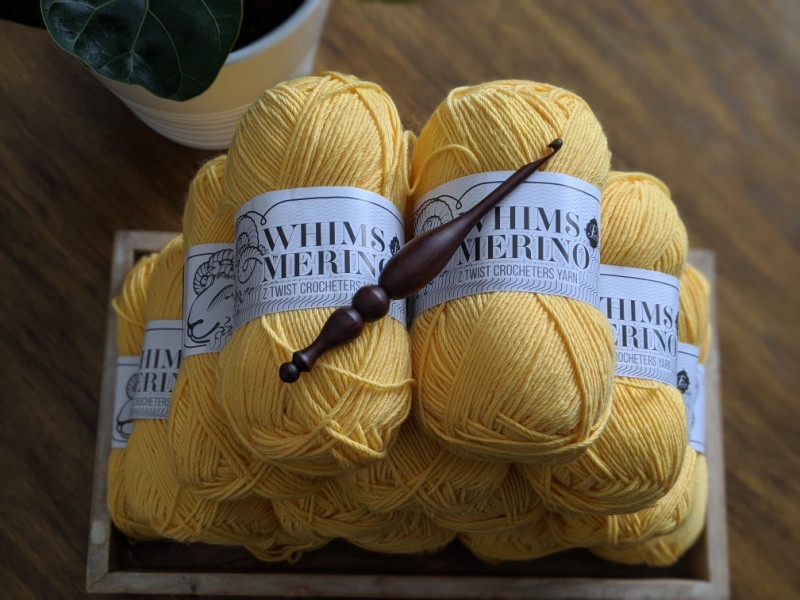 Whims Merino DK in mustard yellow is stacked carefully in a wood tray, with an Alpha Hook placed on top. There is a fiddle leaf fig plant in the top left corner.