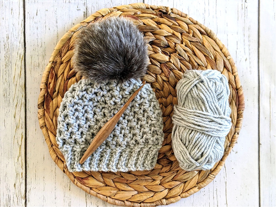 A lansdowne toque in Barva bulky weight yarn is laying on a woven mat. A Furls teak streamline crochet hook lays on top.