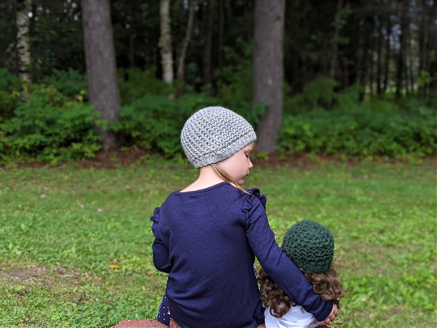 a girl is sitting down outside next to her doll. They are both wearing lansdowne toques.