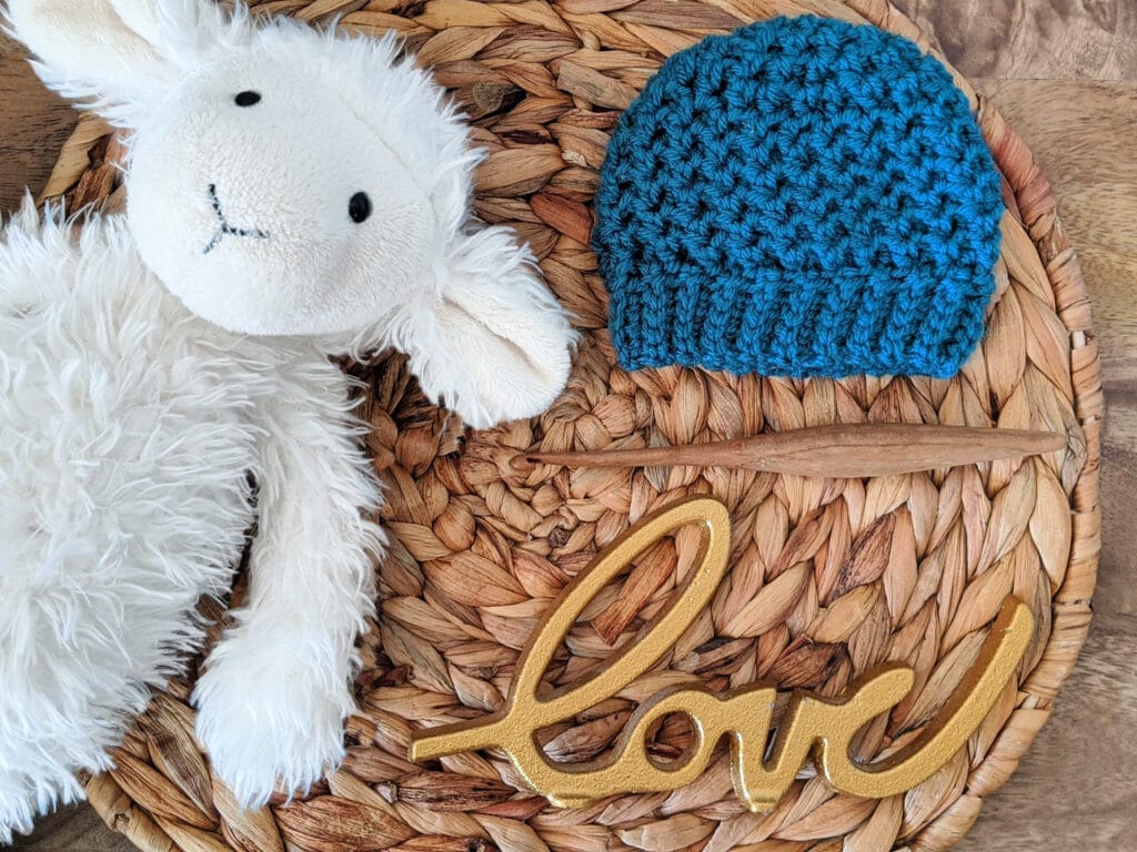 Preemie size Lansdowne Toque in sport weight yarn lays on a round wicker tray beside a stuffed lamb toy and a gold love sign. 