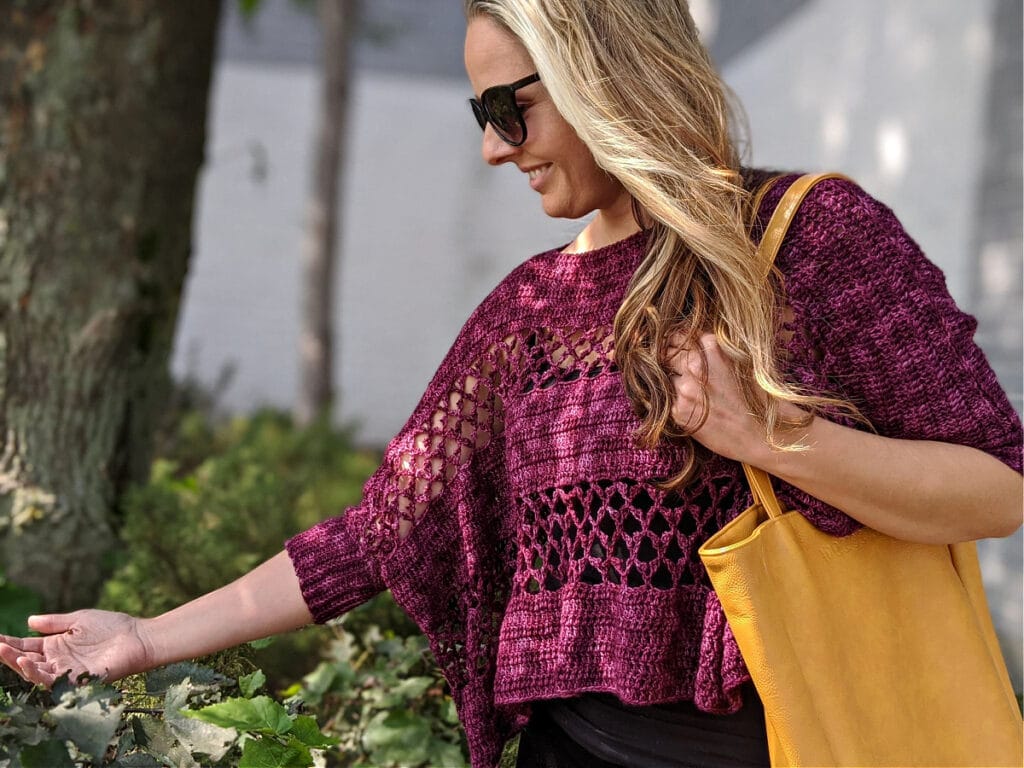 A woman wearing the Contessa Cropped Pullover pattern is touching a leaf, carrying a WeCrochet tote bag.