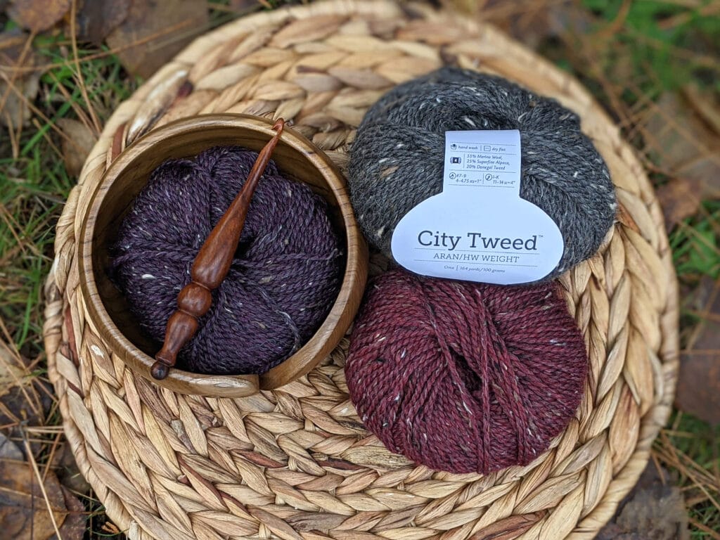 city tweed aran in grey, purple, and maroon are sitting on a wooden circle. A yarn bowl and Alpha series hook from furls crochet lay on top.