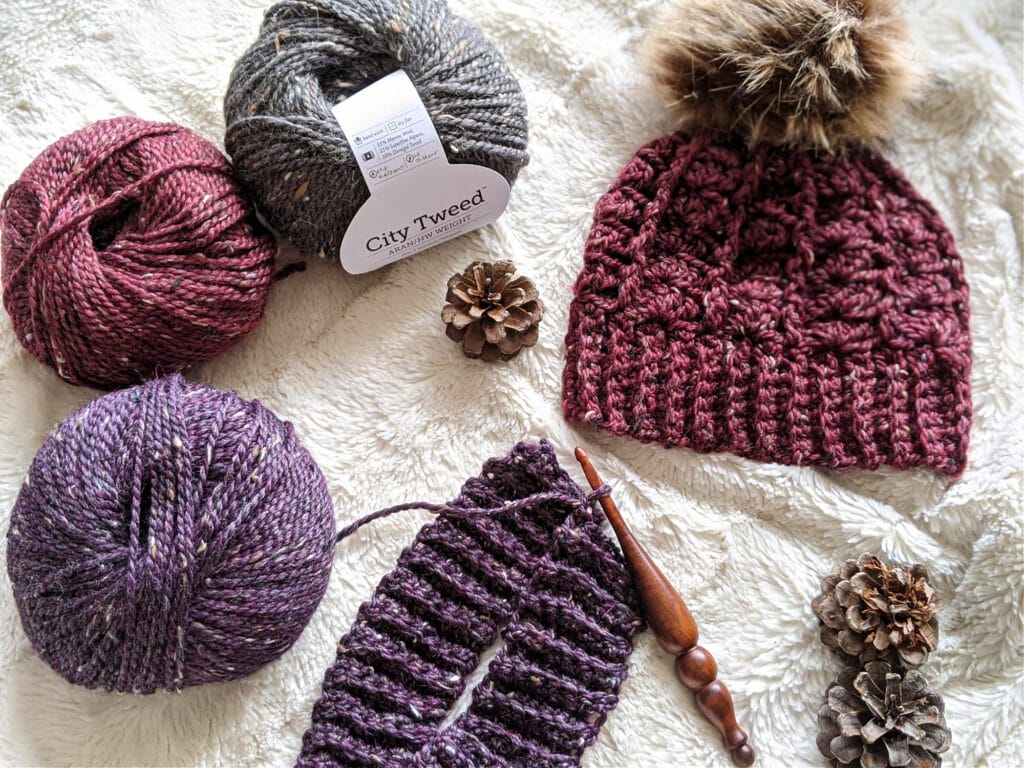 A maroon sarah toque with a fur pompom lays on a white background. An Alpha series crochet is crocheting a second hat in purple. City Tween Aran yarn and pinecones are scattered around.