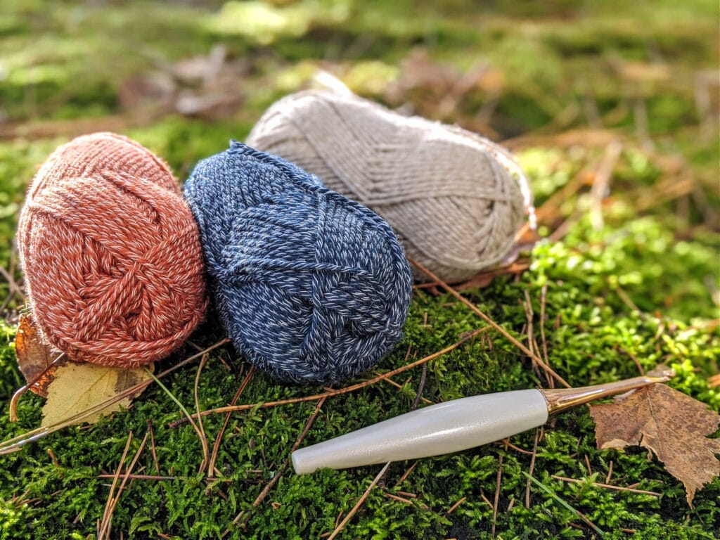 Kindred yarn from WeCrochet is sitting on a bed of moss. A Furls Odyssey crochet hook lays in the foreground.