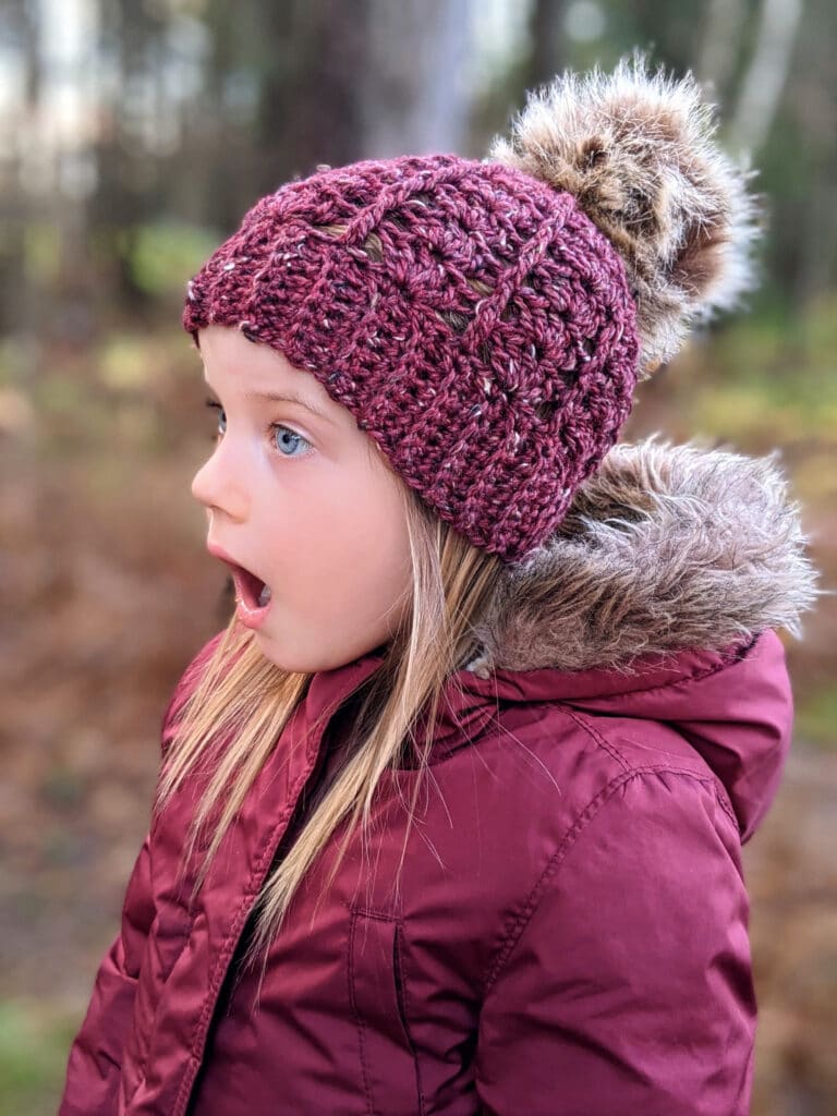 a young girl wears a maroon parka and sarah toque in front of a wooded area.