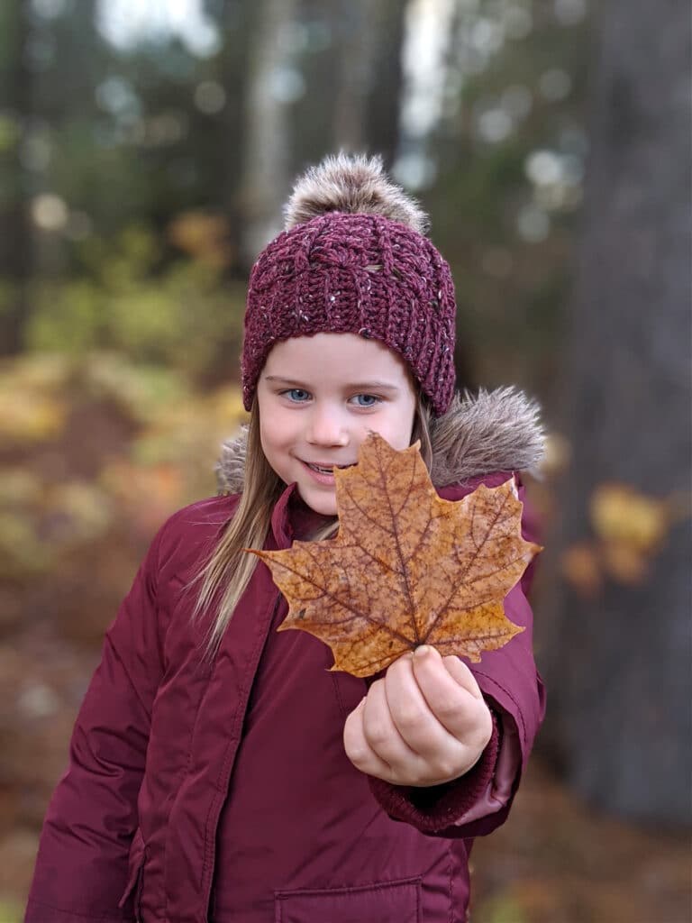 a young girl wears a maroon parka and sarah toque in front of a wooded area.