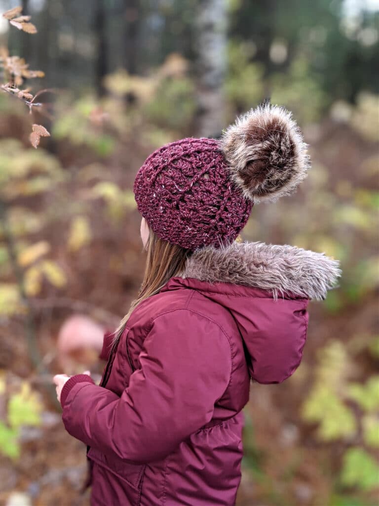 a young girl wears a maroon parka and sarah toque in front of a wooded area.