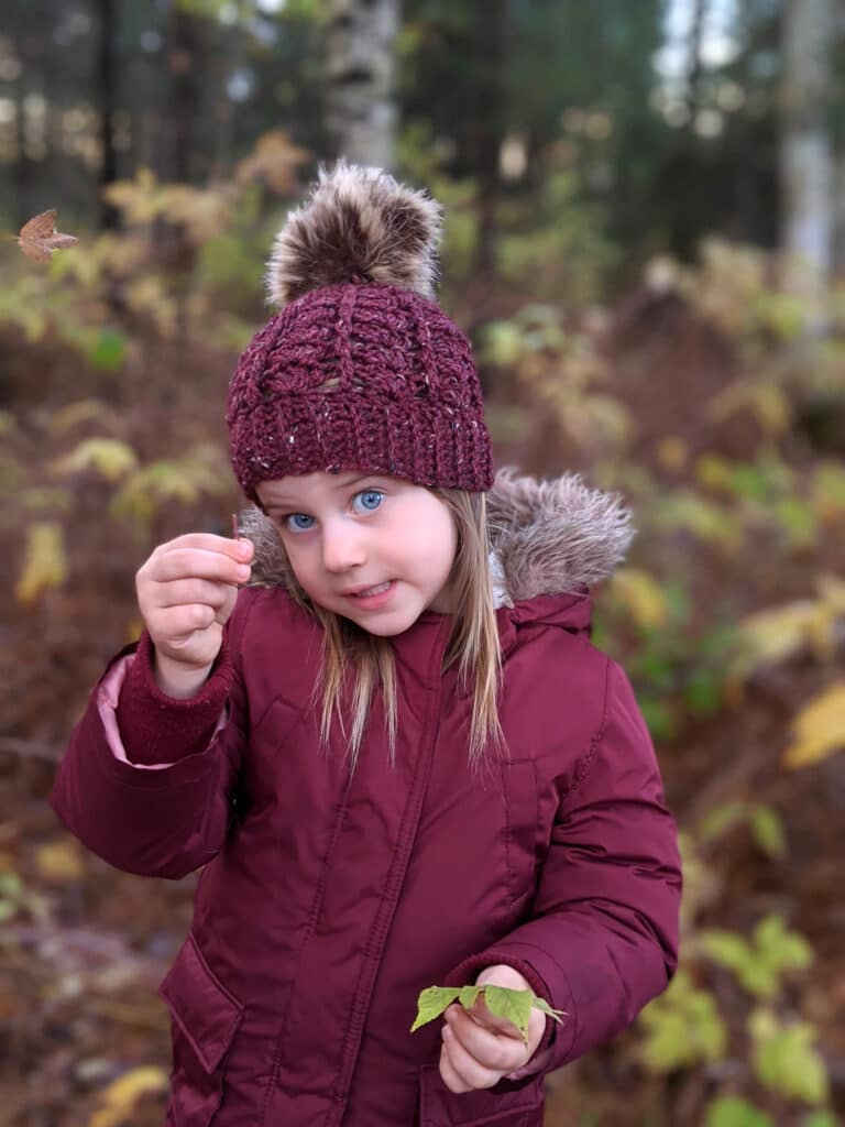 a young girl wears a maroon parka and sarah toque in front of a wooded area.