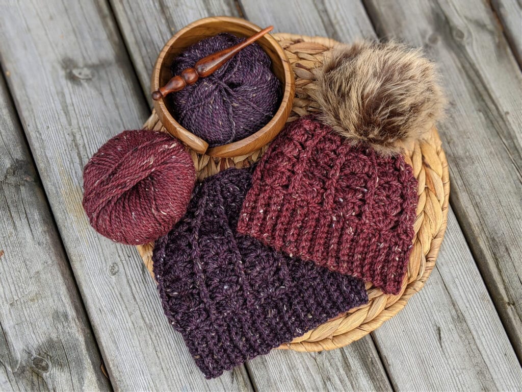 a purple and a maroon sarah toque lay on a wicker mat. City Tweed Aran weight yarn lies in a yarn bowl and on a wood background. An Alpha series crochet hooks lays on top.