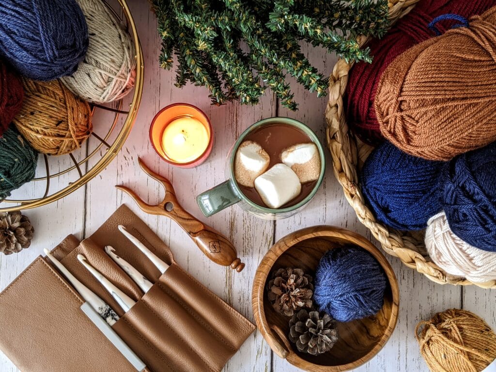 A table top filled with bowls of yarn, crochet hooks, a cup of hot chocolate, and a candle.