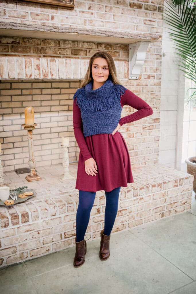 A woman wearing the Bredele Vest crochet pattern is standing in front of a fireplace.