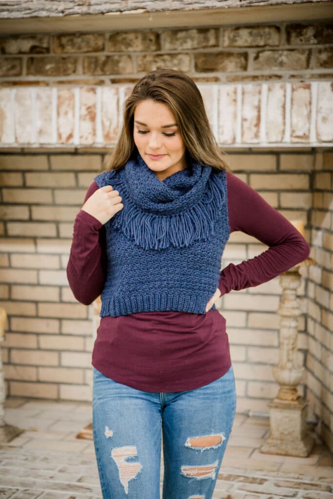 A woman wears Bredele Vest crochet pattern in front of a fireplace.
